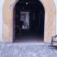 Fotoaufnahme des Haupteingangs vom Gasthaus aus Mühlhausen bzw. vom Wirtshaus am Kommunbrauhaus am aktuellen Standort. Die zweiflügelige Holztür ist nach innen geöffnet, darüber steht „JS 72“. Der Boden vor dem Gebäude ist gepflastert. 