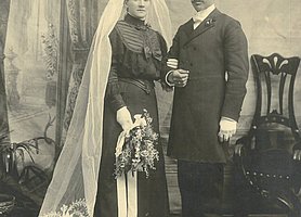 Ehepaar. Die Braut trägt ein schwarzes Kleid. Dazu trägt sie einen weißen Schleier. Der Bräutigam trägt einen schwarzen Anzug.