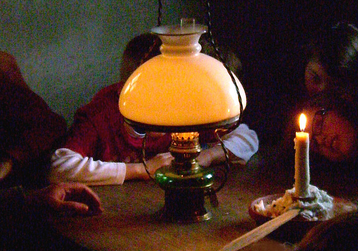 Fotoaufnahme der Führung „Feuer und Licht im Haus“. Um einen Holztisch sitzt eine Gruppe Kinder. In der Mitte stehen eine leuchtende Petroleumlampe und eine angezündete Kerze. Abgesehen von diesen schwachen Lichtquellen ist der Raum dunkel. 