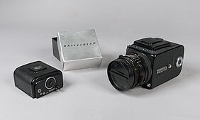 Die Kamera ist schwarz. Sie sieht aus wie eine Würfel. An dem Würfel ist ein rundes Objektiv. Daneben liegt eine kleine schwarze Schachtel. Sie ist ein Filmmagazin. Die Verpackung zum Magazin ist silber.