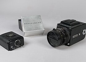 Die Kamera ist schwarz. Sie sieht aus wie eine Würfel. An dem Würfel ist ein rundes Objektiv. Daneben liegt eine kleine schwarze Schachtel. Sie ist ein Filmmagazin. Die Verpackung zum Magazin ist silber.