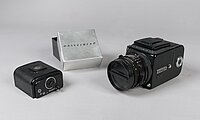 Die Kamera ist schwarz. Sie sieht aus wie eine Würfel. An dem Würfel ist ein rundes Objektiv. Daneben liegt eine kleine schwarze Schachtel. Sie ist ein Filmmagazin. Die Verpackung zum Magazin ist silber.