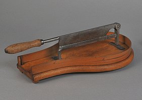 Der Zuckerschneider ist ein Messer. Es sitzt auf einem Holzbrett. Man kann das Messer herunterklappen. Das Brett hat die Form eines Tropfens.