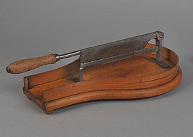 Der Zuckerschneider ist ein Messer. Es sitzt auf einem Holzbrett. Man kann das Messer herunterklappen. Das Brett hat die Form eines Tropfens.