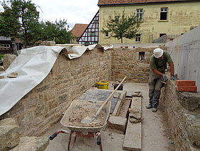 niedrig aufgemauerte Sansteinmauern, darin arbeitet ein Maurer. Schubkarre und Werkzeug umgibt ihn.